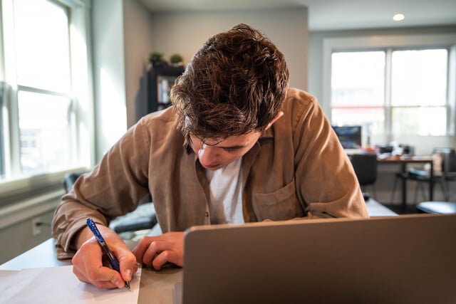 Estudiar a distancia qué es. Chico tomando notas frente a su ordenador mientras estudia a distancia.